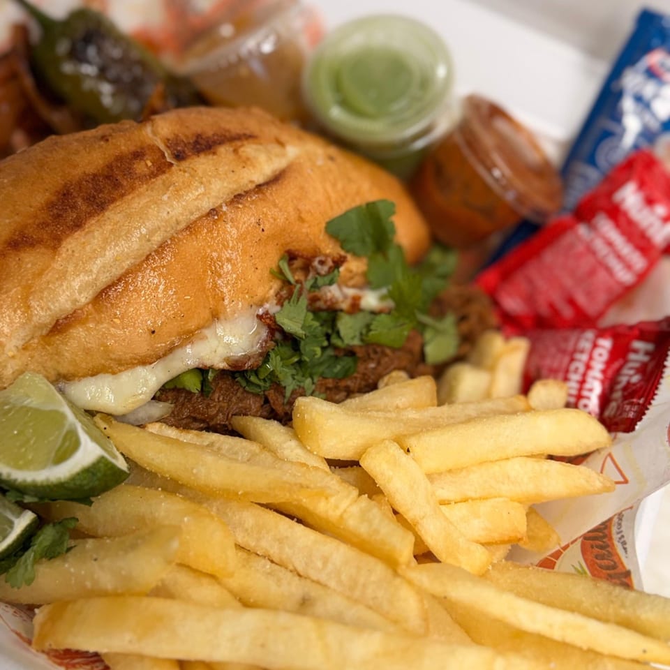 A close-up of a torta sandwich with melted cheese, cilantro, lime, fries, and sauces.