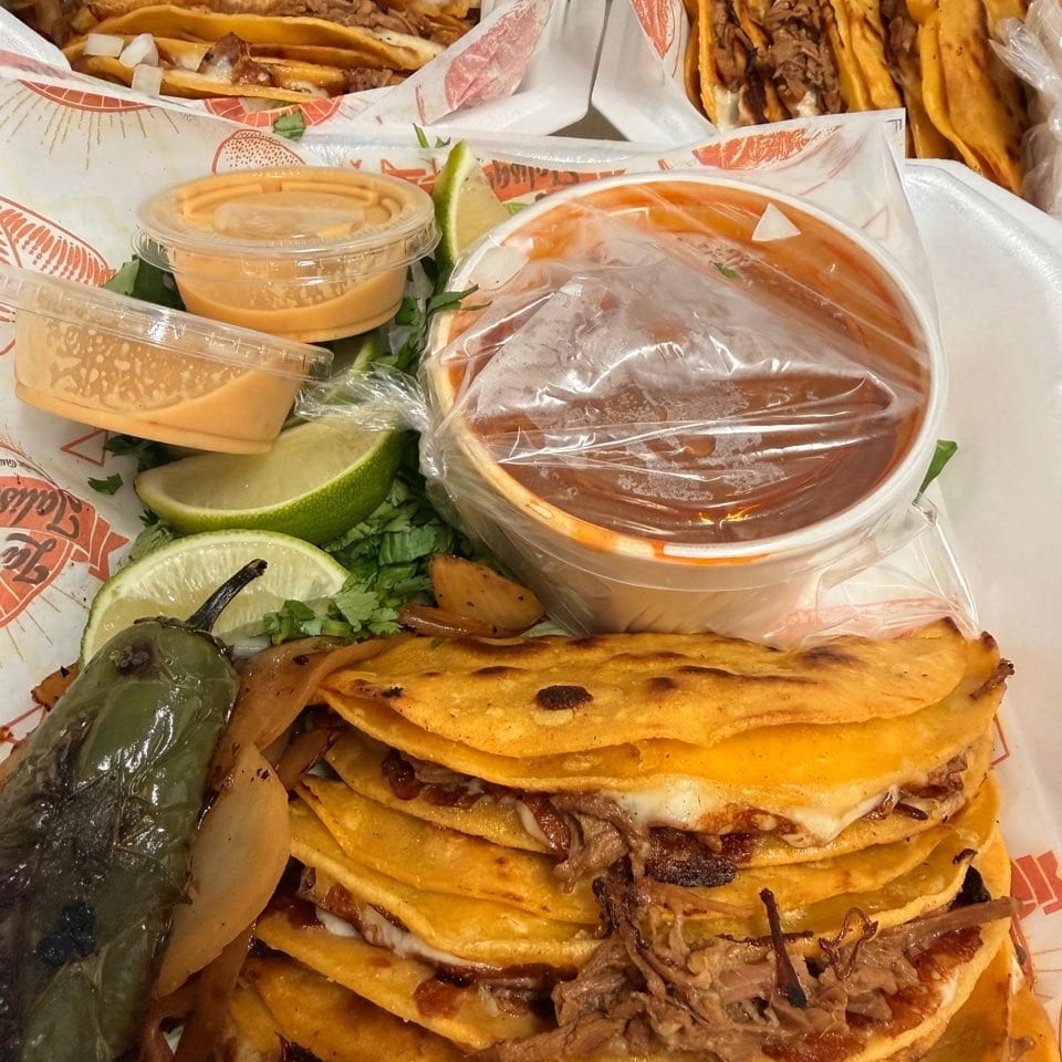 Several birria tacos with limes, sauces, and a roasted pepper on a white plate.