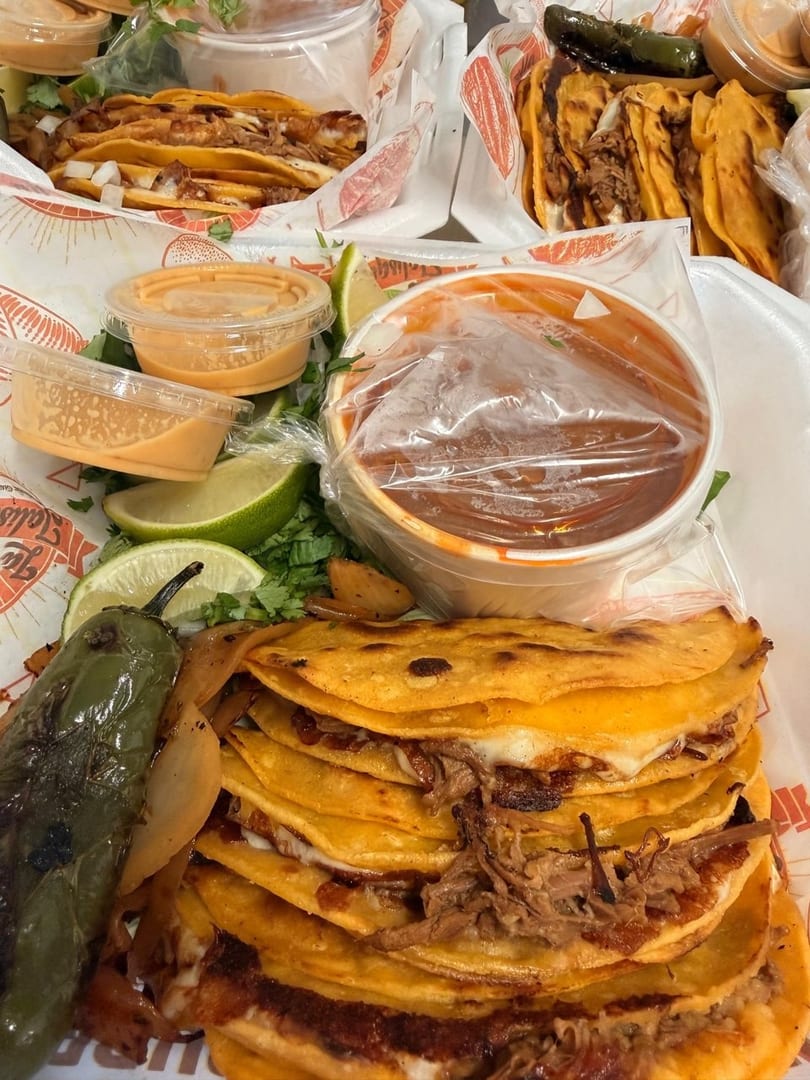Several birria tacos with limes, sauces, and a roasted pepper on a white plate.